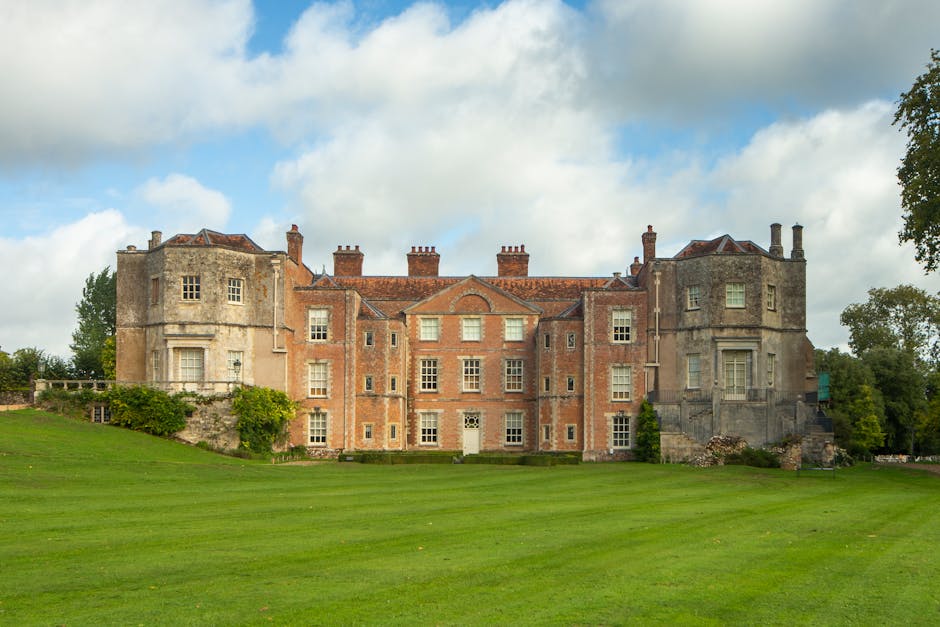 A large, historic stone and brick castle-like building with multiple towers and chimneys, situated on a well-maintained green lawn. The building features a combination of grey stone walls and reddish-